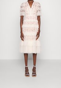 Šaty s více vrstvami délky ke kolenům v světle krémové barvě, s průsvitnými nadýchanými rukávy, květinovou výšivkou a texturovanými puntíky. Doplněné sandály na klínku se šňůrkami.