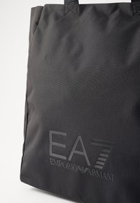 Juodas tosmaišis, pagamintas iš tekstūruotos medžiagos, su dideliu iškilniu логотипu "EA7 EMPORIO ARMANI" priekyje, su dviem tvirtais rankenomis.