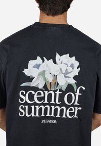 Zwart katoenen t-shirt met een grote bloemenafbeelding met witte bloemen en groene bladeren, naast de tekst "geur van de zomer" in vetgedrukte witte letters.