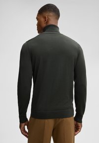 Ein Mann steht mit dem Rücken zur Kamera, trägt einen dunkelgrünen Reißverschluss-Pullover und beigem Hose vor einem hellgrauen Hintergrund.