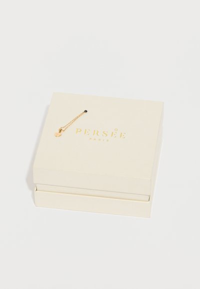 Caixa de joias de cor creme com texto dourado, contendo uma delicada corrente dourada com um pequeno detalhe preto. Acabamento suave, design minimalista.