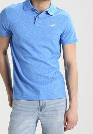 Mann trägt ein blaues kurzärmeliges Poloshirt mit Kragen und hellen Jeans, steht mit einem Arm angewinkelt und dem anderen entspannt.