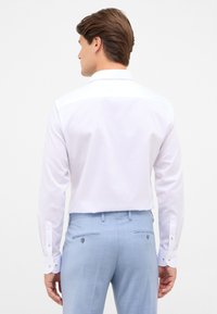 Camicia bianca a maniche lunghe con colletto classico, dotata di polsini con bottoni. Abbinata a pantaloni azzurri chiari che presentano un tessuto texture.