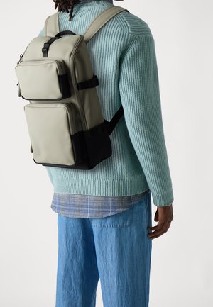 Rucksack aus hellgrauem Material mit mehreren Taschen, schwarzen Akzenten, verstellbaren Trägern und einem schlanken Design; getragen über einem strukturierten blauen Pullover.