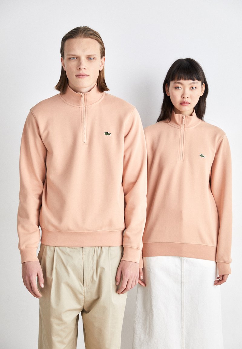 Felpe quarter-zip rosa chiaro con polsini a coste e orli arrotondati, caratterizzate da un piccolo logo verde sul petto. Un modello indossa dei pantaloni beige, l'altro una gonna bianca.
