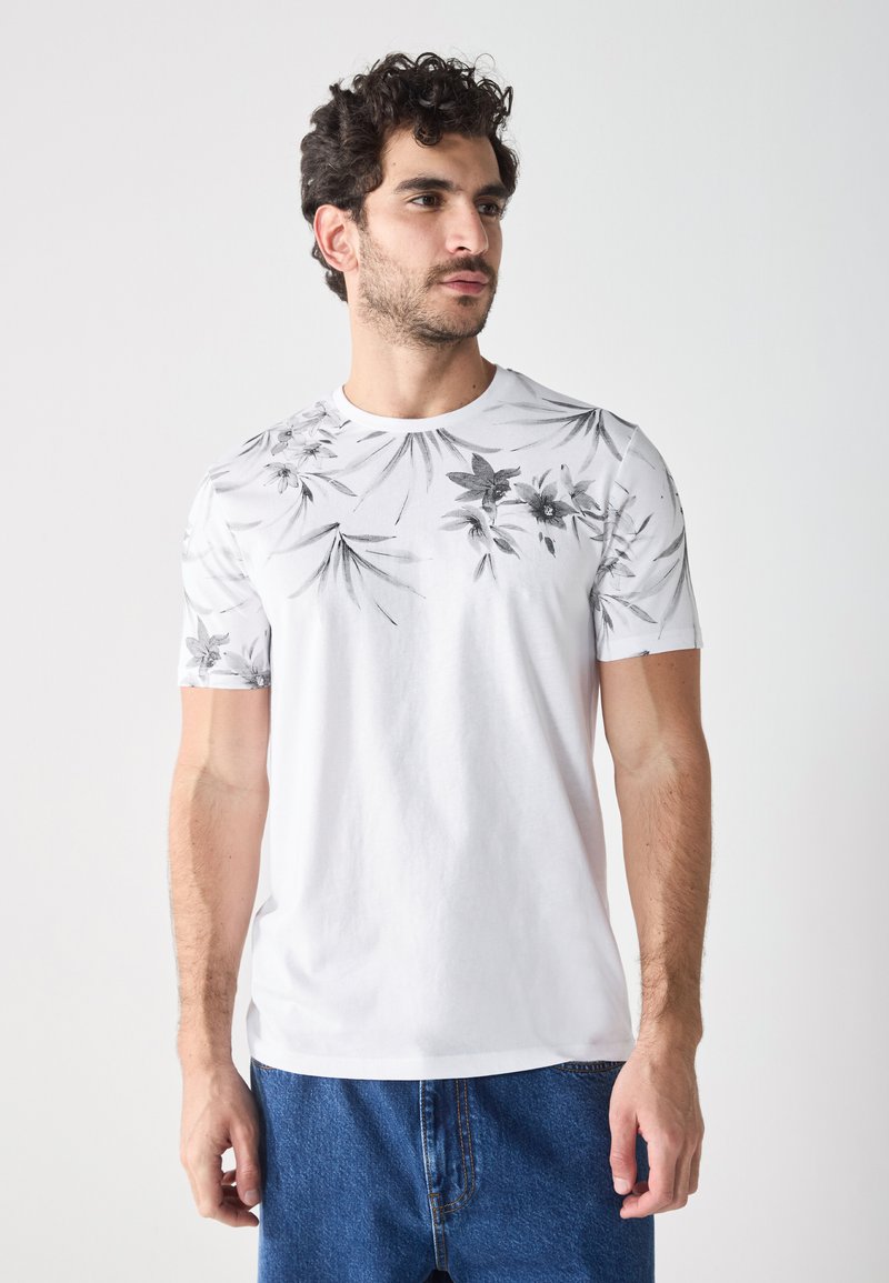 White cotton t-shirt featuring a black floral print on short sleeves and upper body. Crew neckline, relaxed fit, paired with blue denim shorts.
