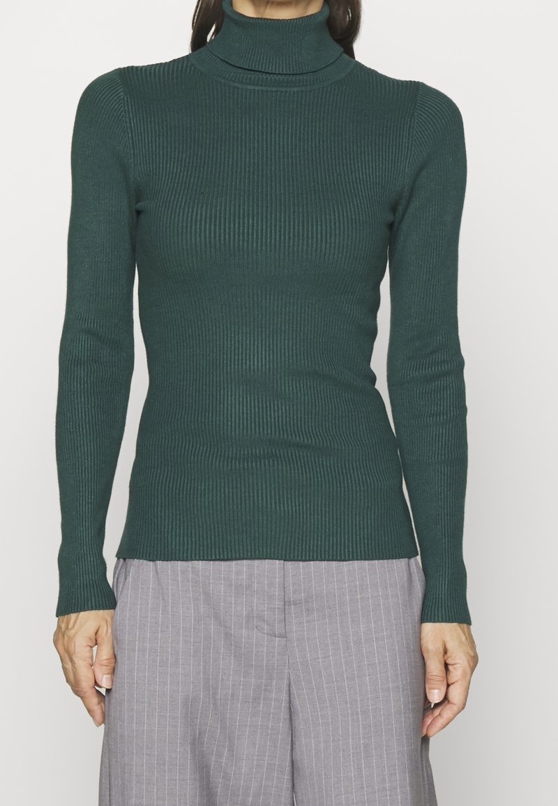 Pull à col roulé côtelé vert foncé avec manches longues, design ajusté, texture douce et ourlet droit, associé à un pantalon gris clair à rayures.