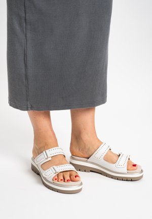 Füße in weißen Sandalen mit zwei Riemen und goldenen Nieten, dazu rote Fußnägellackierung, kombiniert mit einem grauen, gerippten Midirock vor weißem Hintergrund.