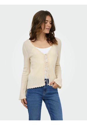 Femme portant un cardigan en maille côtelée crème avec des bords festonnés et des liens, sur un top blanc et un jean bleu, posant avec une main dans la poche.