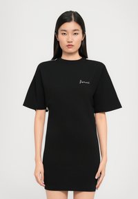 Γυναίκα με μακριά μαύρα μαλλιά στέκεται μπροστά φορώντας ένα μαύρο κοντομάνικο φόρεμα με το λογότυπο "fiorucci" στο στήθος.