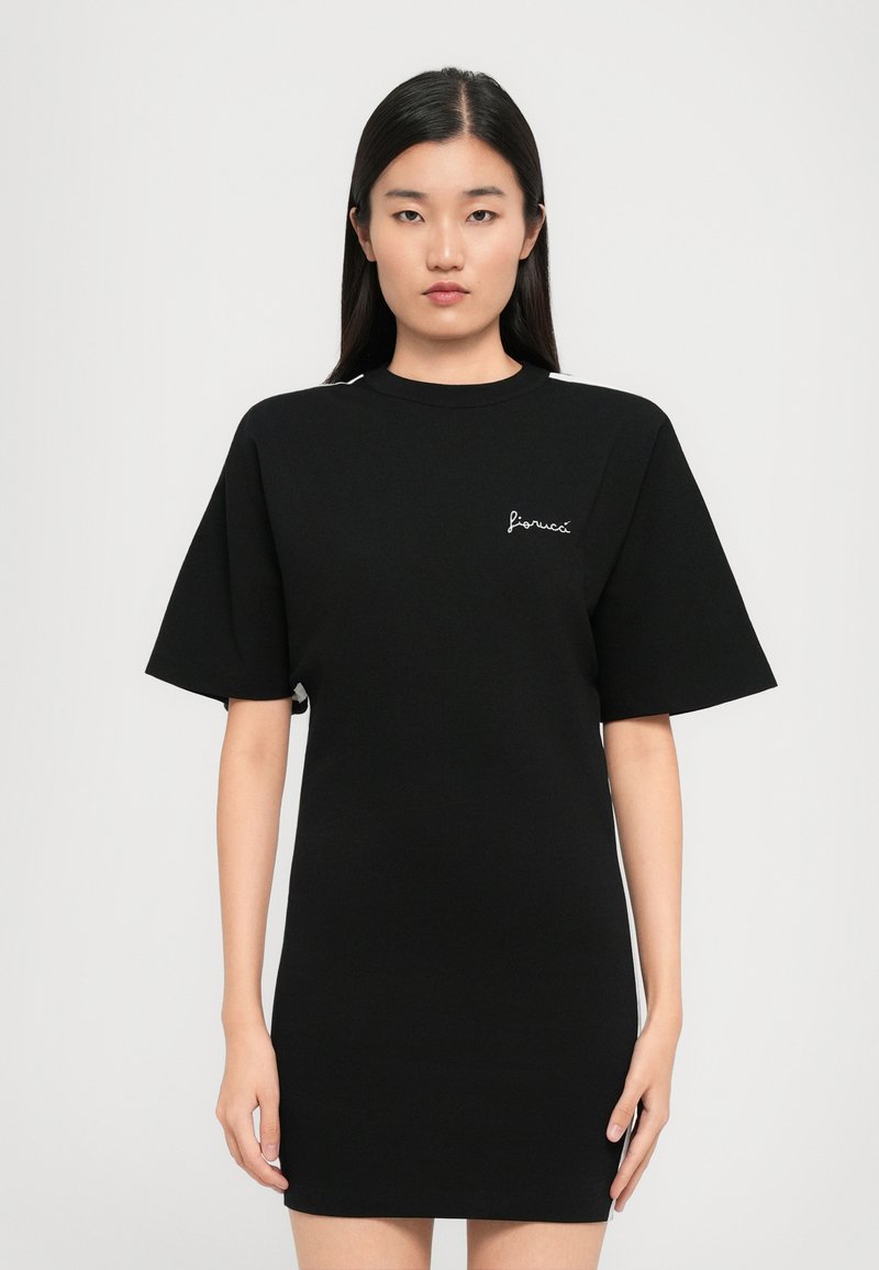 Γυναίκα με μακριά μαύρα μαλλιά στέκεται μπροστά φορώντας ένα μαύρο κοντομάνικο φόρεμα με το λογότυπο "fiorucci" στο στήθος.