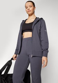Tamno siva zip-up dukserica s teksturiranim završetkom, u kombinaciji s crnom crop majicom i odgovarajućim suženim trenirkama. Drži crnu torbu.