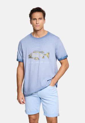 Hombre con camiseta azul claro con gráfico de pez y pantalones cortos azul claro remangados, de pie con una mano en el bolsillo contra un fondo blanco.