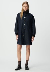 Czarna sukienka koszulowa z długimi rękawami, zapinana na guziki z przodu, z jedną kieszenią na piersi i subtelnym połyskiem. Posiada złote guziki i swobodny krój.