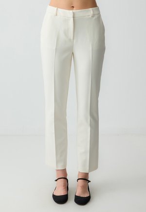 White tailored pants with a smooth fabric, flat front, and side pockets. Cropped length, straight legs, worn with black flats.