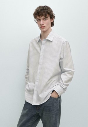 Junger Mann mit lockigem braunem Haar trägt ein hellgraues, dünn gestreiftes Hemd mit Knöpfen und blaue Jeans, steht mit den Händen in den Taschen.