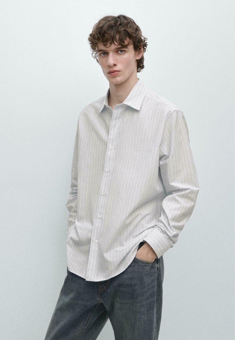 Jeune homme aux cheveux bruns bouclés portant une chemise boutonnée gris clair à fines rayures et un jean bleu, debout les mains dans les poches.