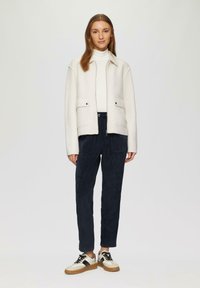 Lichtgekleurde wollen jas met een kraag, twee voorkruiden, gecombineerd met donkere broek en sneakers met een zwart-wit ontwerp.