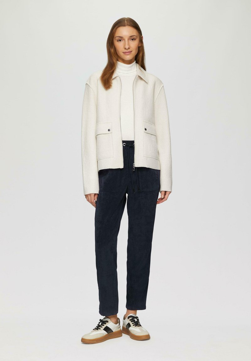 Veste en laine de couleur claire avec col, deux poches avant, associée à un pantalon sombre et des baskets au design blanc et noir.