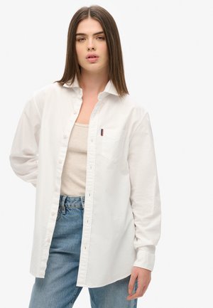 Biała koszula na guziki z bawełny z kieszonką na piersi, kołnierzykowym wzorem i długimi rękawami, noszona na beżowym topie bez rękawów.