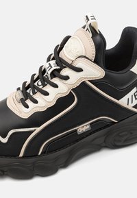 Schwarze und cremefarbene Sneaker mit einer strukturierten Lederoberfläche, gepolstertem Kragen und einer robusten Gummisohle mit kreisförmigen Profilmustern. Hardware-Logo-Detail.