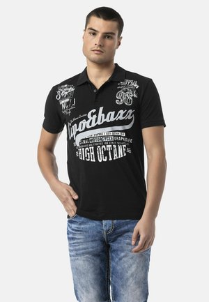 Jeune homme portant un polo noir avec un texte blanc et un jean bleu, debout avec une main dans la poche sur un fond uni.
