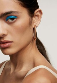 Pendientes de aro en oro rosa con un acabado pulido, que presentan una gran forma circular y una textura suave, llevados por un modelo con un maquillaje mínimo.