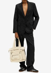 Schwarzer Blazer mit floralem Muster, kombiniert mit schwarzen Cargo-Hosen und einer beigefarbenen Tragetasche. Schwarze Sneaker mit weißen Akzenten runden den Look ab.