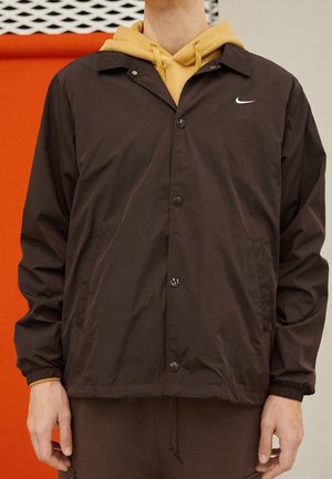 Brown windbreaker jacket with a button-up front, featuring a small white logo, and elasticized cuffs. Worn over a yellow hoodie.