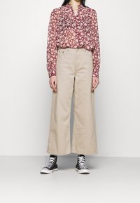 Blusa floreale con scollatura arricciata in rosso e crema; abbinata a pantaloni a gamba larga beige. Sneakers alte nere completano l'outfit.