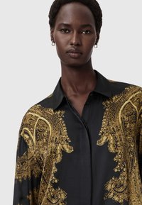 Camisa preta com intrincado padrão paisley dourado, mangas longas e colarinho clássico. O tecido tem uma textura suave e brilhante.