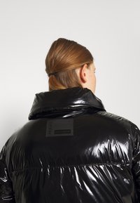 Sort pufferjakke med en blank finish, høj krave og en rektangulær mærkat på bagsiden. Har syede detaljer og synlige sømme.