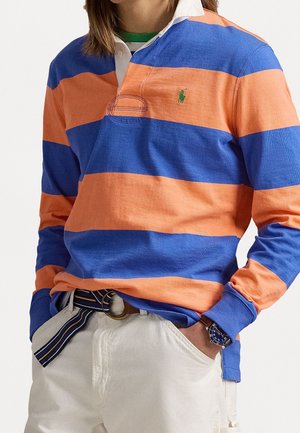 Koszula polo z długim rękawem w pomarańczowo-niebieskie poziome paski, z białym kołnierzykiem i zielonym logo akcentującym. Nosić z beżowymi spodniami i paskiem w paski.