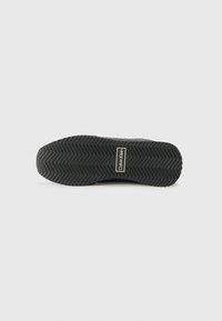 Svart sneakersåle med strukturert sikksakk mønster, med et logo-patch i hvitt og gull, laget av gummimateriale.
