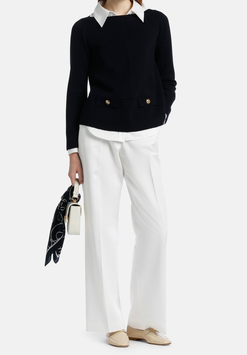 Femme portant un pull noir par-dessus une chemise blanche à col, pantalon blanc à larges jambes, chaussures plates beiges, tenant un sac à main blanc avec une écharpe noire.