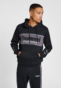 Svart hoodie med en grå panel som visar "hummel SPORTSWEAR"-logotypen. Inkluderar dragsnören, känguruficka och chevrondetaljer på ärmarna.