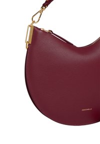 Borsa in pelle bordeaux a forma di mezzaluna, con hardware dorato e dettaglio del logo scolpito sul fondo. Texture liscia con chiusura a zip.