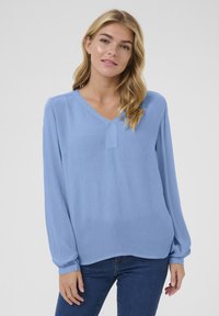 Blouse à manches longues bleu clair avec un col en V, en tissu doux présentant une texture subtile. Le design comprend une couture centrale et une coupe décontractée.