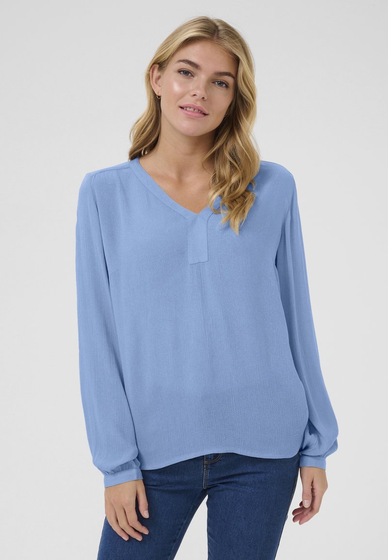 Blouse à manches longues bleu clair avec un col en V, en tissu doux présentant une texture subtile. Le design comprend une couture centrale et une coupe décontractée.