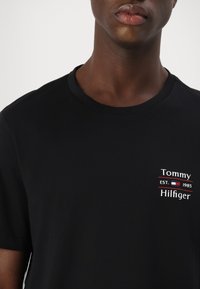Black cotton T-shirt featuring a crew neck, short sleeves, and a white and red printed logo with "Tommy Hilfiger" and establishment year.