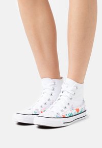 Białe płócienne sneakersy typu high-top z kwiatowym wzorem po bokach, czarna gumowa podeszwa i metalowe oczka na sznurowadła.