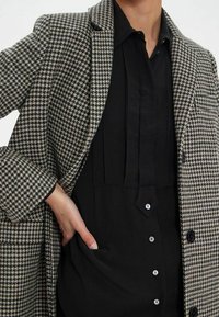 Manteau à motif pied-de-poule en tissu gris et noir, présentant un design à double boutonnage et une chemise noire contrastante avec des plis.