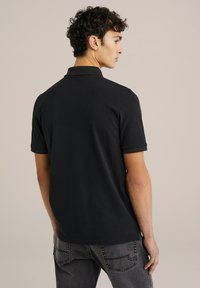 Musta polo-paita pehmeällä pinnalla, lyhyet hihat, klassinen kaulus ja sivureiät, yhdistettynä harmaisiin farkkuihin, nähtynä takaa.