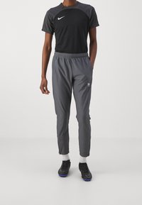 Schwarzes Sportshirt mit strukturierten Ärmeln, graue Sporthose mit elastischem Bund und Logo, kombiniert mit schwarzen Sportschuhen und weißen Socken.