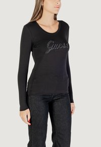 Top lungo nero a maniche lunghe con scollo rotondo, caratterizzato dal logo "Guess" decorato con accenti brillanti. Abbinato a jeans in denim scuro.