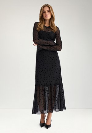 Femme debout, bras croisés, portant une longue robe noire transparente à pois et des escarpins noirs pointus sur un fond clair uni.