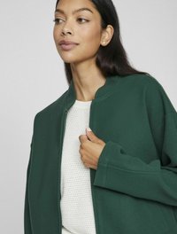 Grön zip-up jacka med rund halsringning, tillverkad av texturerat tyg; lagrad över en vit, stickad tröja.
