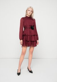 Γυναίκα που φοράει ένα μακρυμάνικο,  tiered μπορντό φόρεμα με μαύρη λουλουδάτη ζώνη, στέκεται μπροστά σε λευκό φόντο.