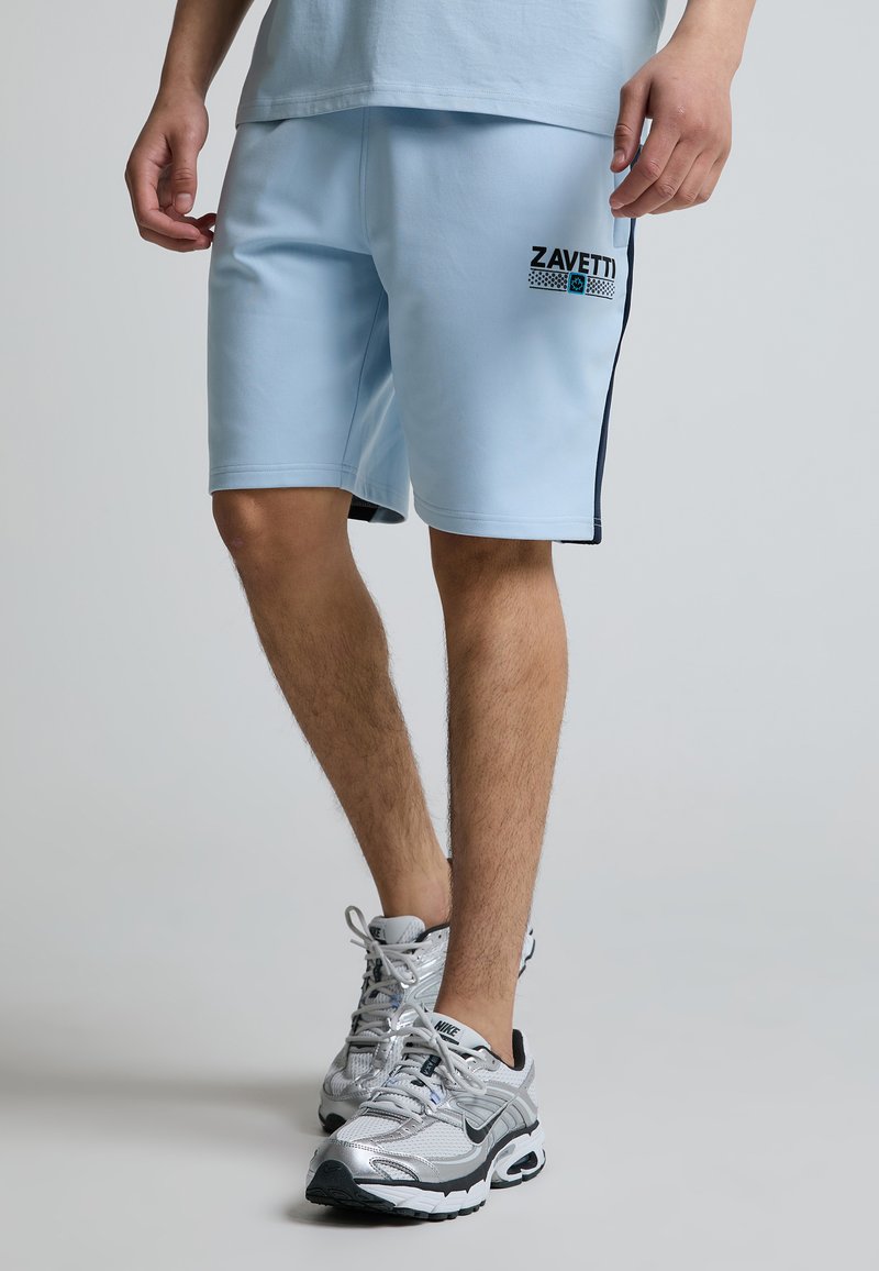 Jambes masculines portant un short Zavetti bleu clair et des chaussures de running Nike argentées, debout sur un fond uni gris clair.