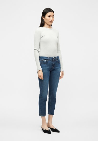 Femme debout portant un haut à manches longues gris clair, un jean court bleu et des ballerines à bride arrière pointues noires, sur un fond blanc uni.